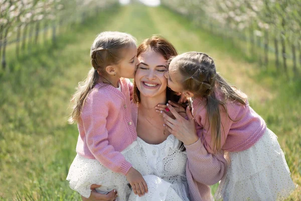 Портрет матері та її дочок-близнючок у квітучому саду яблук. Дівчата цілують маму. — стокове фото