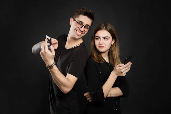 Portrait of a young couple standing with mobile phone, man is playing games on mobile phone while angry girl standing near and looking at camera isolated over black background.