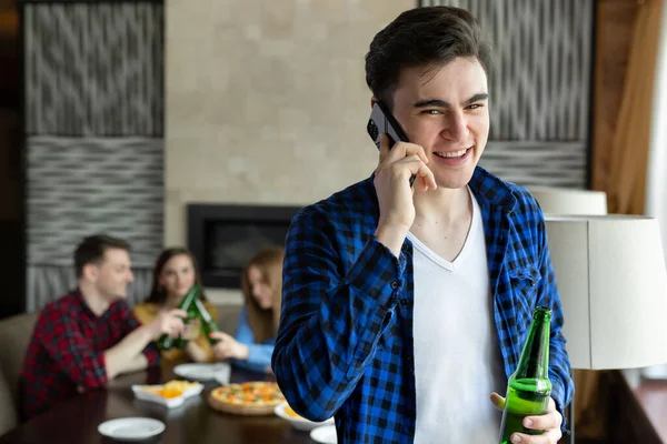 Jovem bebe cerveja de uma garrafa, fala ao telefone e olha pela janela de um café contra o fundo de seus amigos que estão sentados à mesa e comendo pizza — Fotografia de Stock