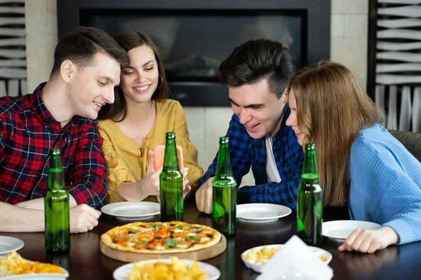 Счастливые друзья смотрят в телефон, когда пьют пиво и пиццу в кафе. — стоковое фото