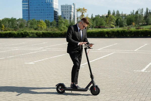 Joven hombre de negocios guapo en un traje monta una scooter eléctrico y mira a un ordenador portátil — Foto de Stock