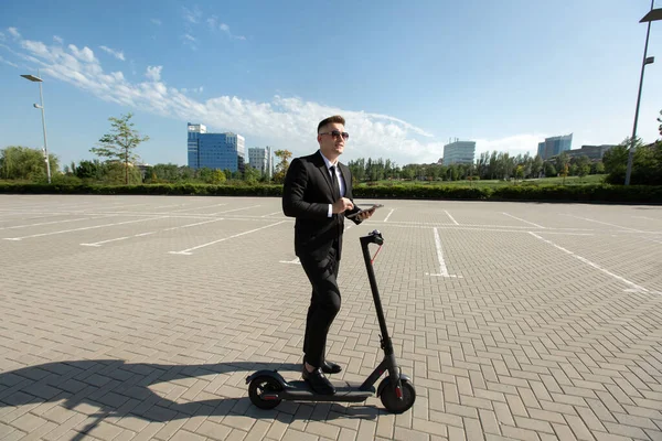 Joven hombre de negocios guapo en un traje monta una scooter eléctrico y mira a un ordenador portátil —  Fotos de Stock