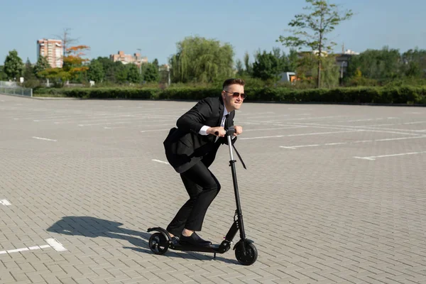 Joven hombre de negocios guapo en un traje monta un scooter eléctrico alrededor de la ciudad y se ríe — Foto de Stock