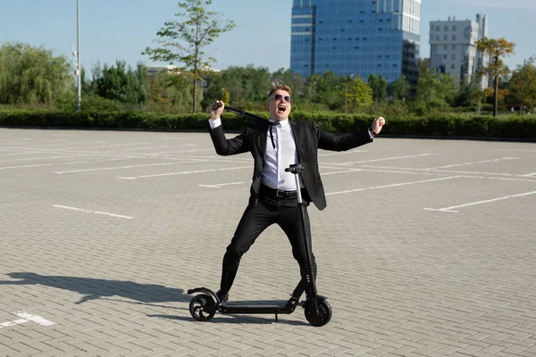 Joven hombre de negocios guapo en un traje monta un scooter eléctrico alrededor de la ciudad y se ríe — Foto de Stock