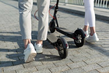 Bir çift kadın ve erkek parkta yürüyorlar ve elektrikli scooter sürüyorlar.