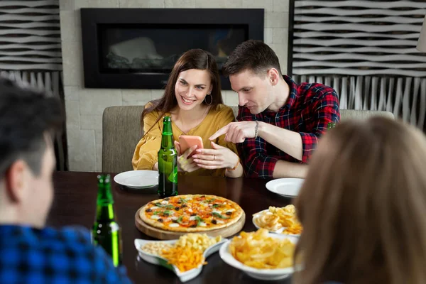 Grupo de amigos em um café com pizza e cerveja se divertindo. A menina mostra ao cara uma foto engraçada no telefone — Fotografia de Stock