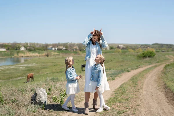Anne ve ikiz kızları ineklerin yakınındaki bir köyde toprak bir yol boyunca yürüyorlar. Anne ve kızları inek gibi davranıp eğleniyorlar. — Stok fotoğraf
