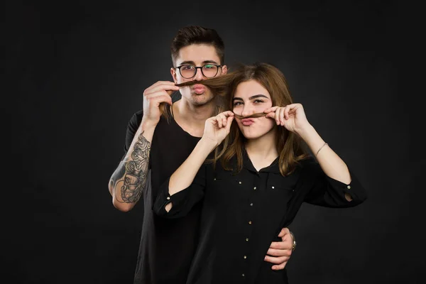 Bela jovem casal amoroso fazendo bigode falso do cabelo, enquanto em pé contra fundo preto. bigode engraçado — Fotografia de Stock