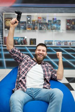 Genç, mutlu ve heyecanlı bir adam elinde bir kontrolörle bir sandalyeye oturur ve video oyunları oynar ve eğlenir. VR, konsol oyun alanı.