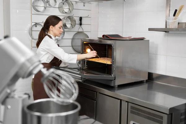 La pasticciera donna mette la pasta di spugna in forno e controlla l'impasto con un bastoncino di legno, uno spiedino — Foto Stock