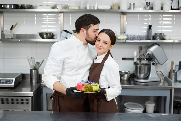 Felice coppia amorevole di pasticceri, un uomo e una donna tenere una torta di mousse nelle loro mani in una cucina professionale — Foto Stock