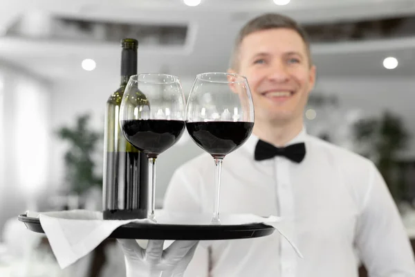 Garçom masculino sorridente com copos de vinho tinto e uma garrafa em uma bandeja em um restaurante — Fotografia de Stock