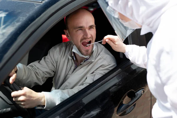 Um homem usando um terno de proteção, luvas, máscara cirúrgica e máscara facial está testando o coronavírus co id-19 em outro homem sentado em um carro — Fotografia de Stock