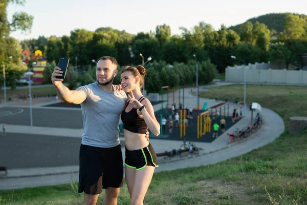 若い幸せな男と女は、スポーツフィールドの背景に公園で電話で自撮りを取ります. — ストック写真