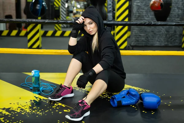 Young athletic girl in a black tracksuit sits in the ring and rests after a workout — Stock Photo, Image