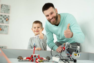 Mutlu yakışıklı adam, lego robotlarla oynarken neşeli küçük oğluyla kameraya gülümsüyor ve baş parmağını gösteriyor.