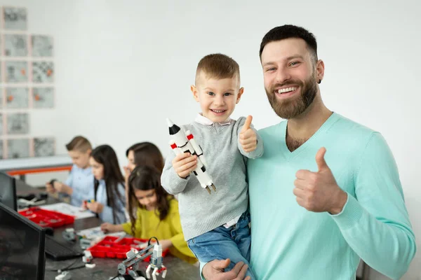 Porträt eines jungen Vaters mit seinem Sohn auf dem Arm zeigt den Daumen nach oben in einem Robotikkurs — Stockfoto