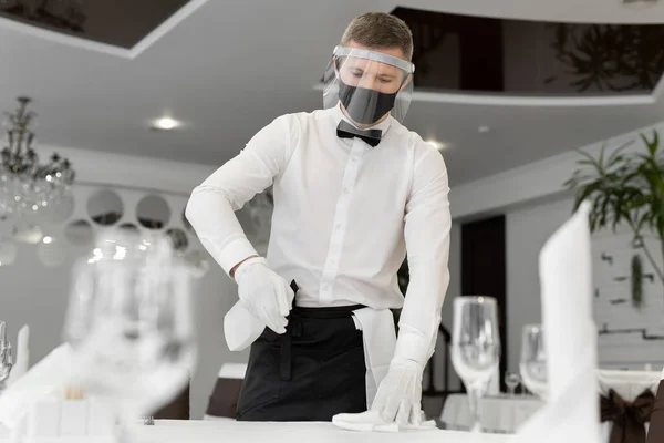 얼굴 마스크와 얼굴 가리개를 착용 한 남성 웨이터의 모습, 사교적 인 거리를 두고 카페에서 탁자를 청소하는 모습. — 스톡 사진