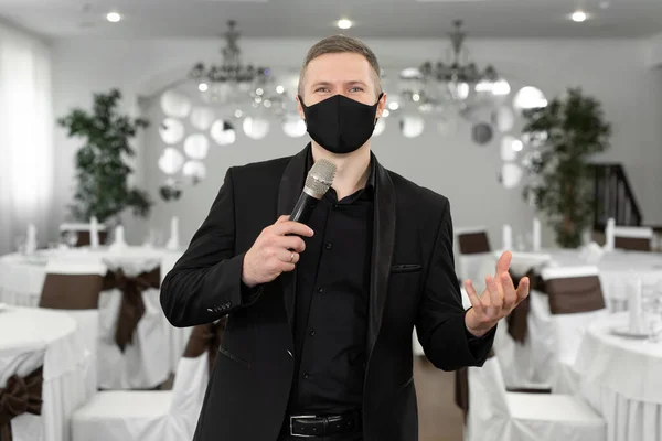 Showman in giacca e cravatta e maschera protettiva con un microfono in mano nella sala banchetti di rastoran — Foto Stock