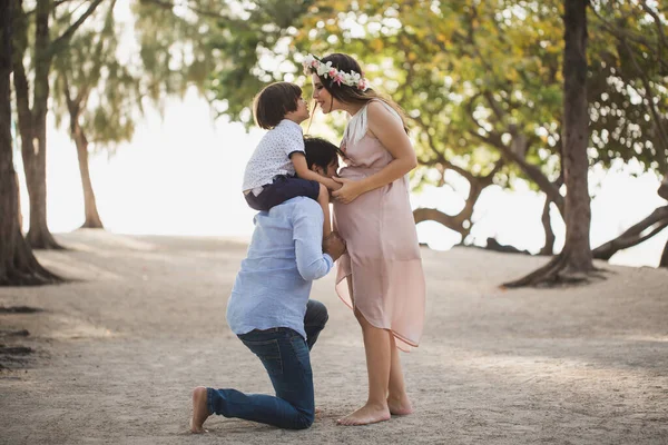 Boldog terhes család szórakozik a természetben. Fiam, az apád vállán. Apa hasba csókolja a terhes feleségét. — Stock Fotó