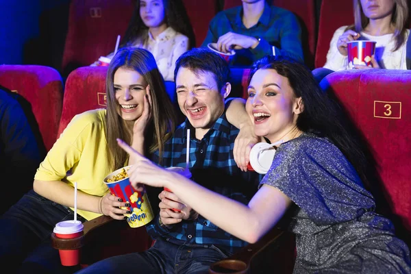 Две молодые девушки и парень, смотрящий комедию в кино. Молодые друзья смотрят кино в кино. Группа людей в театре с попкорном и напитками . — стоковое фото