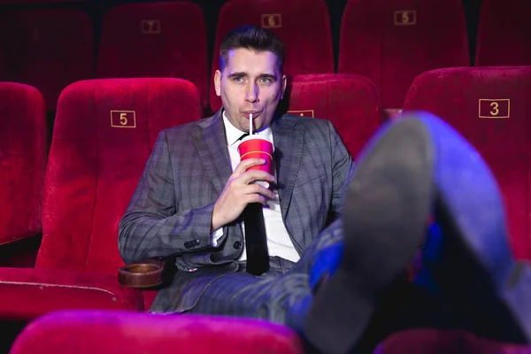 ビジネススーツ姿で映画館に一人でいる若いハンサムな男の肖像で、前の席に足をつけ、赤いカップからチューブを通して飲む — ストック写真
