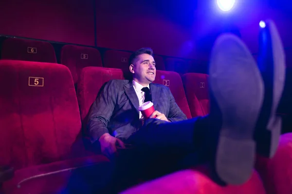 ビジネススーツ姿で映画館に一人でいる若いハンサムな男の肖像で、前の席に足をつけ、赤いカップからチューブを通して飲む — ストック写真
