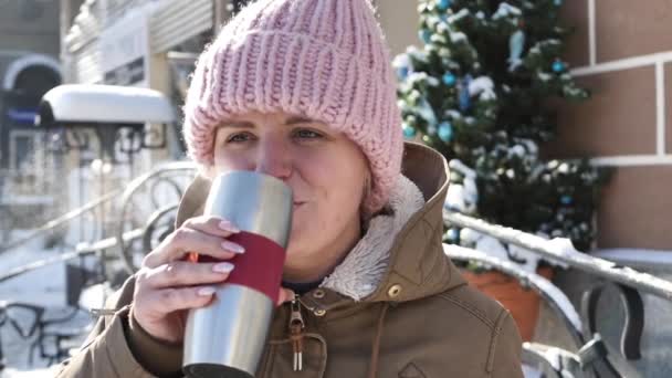 Mladá krásná dívka v teplém klobouku pije čaj z termoup na ulici a usmívá — Stock video