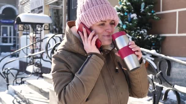 Mladá krásná dívka v teplé čepici pije čaj z termosky na ulici a mluví na mobilním telefonu — Stock video