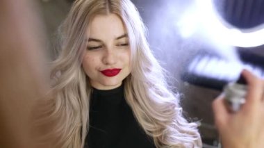 Genç, güzel bir kadın kuaförde oturmuş aynaya bakıyor kuaförü de saç spreyiyle saçını yaptırıyor..