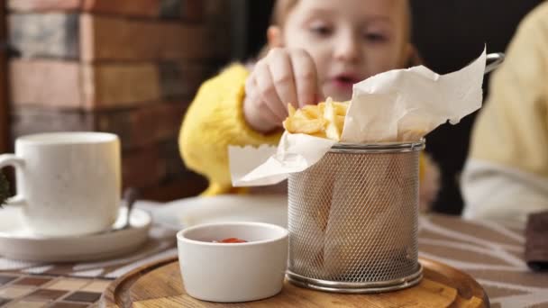 A little girl is eating French fries in a cafe. The child makes faces — Stock Video