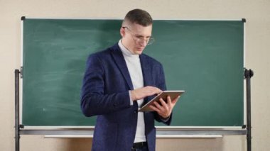 Erkek öğretmen elinde tablet bilgisayar tutarken boş yeşil bir karatahtaya fotokopi alanı ile yazı yazıyor..