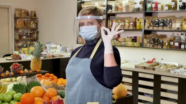 年轻的女保健食品商店店员戴着手套和保护面罩，看着相机，并显示出"还好"的标志 — 图库视频影像