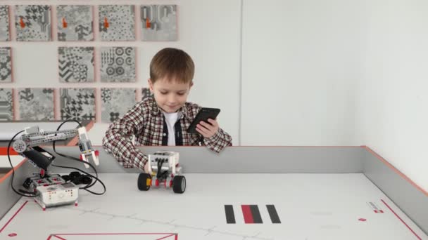 Küçük çocuk robot sınıfında telefonunu kullanarak bir robotu kontrol ediyor. — Stok video