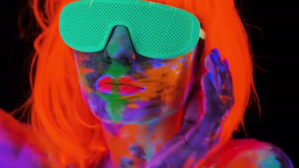 Hermosa mujer modelo en peluca y gafas con colorido maquillaje fluorescente brillante en luz de neón, discoteca — Vídeo de stock