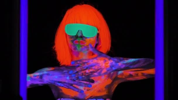 Hermosa mujer modelo en peluca y gafas con colorido maquillaje fluorescente brillante en luz de neón, discoteca — Vídeo de stock