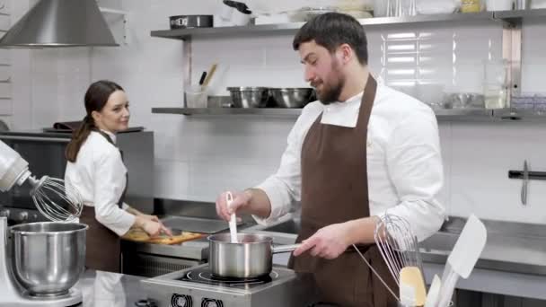 Två bakverk kockar, en man och en kvinna, tillaga dessert, titta på varandra och le. Kärlek i ett professionellt kök — Stockvideo