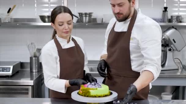 Két cukrász, egy férfi és egy nő, tortát csavar egy standon egy professzionális konyhában. — Stock videók