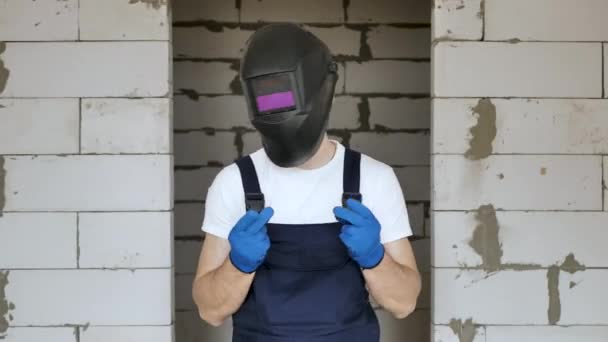 Um trabalhador da construção masculina em um capacete de solda e luvas azuis está dançando e se divertindo em um canteiro de obras — Vídeo de Stock