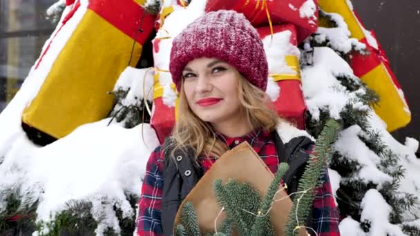 Молодая женщина в красной шляпе с букетом ветвей елки в бумаге во время снегопада — стоковое видео