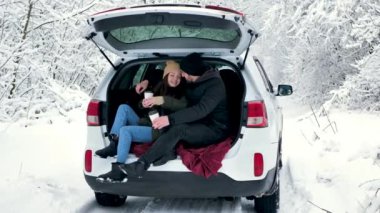 Genç bir çift, bir erkek ve bir kadın, kışın bir arabanın bagajında oturuyorlar, karlı bir ormanda, sarılıyorlar, öpüşüyorlar ve kahve içiyorlar.