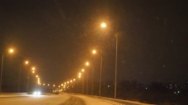 Geceleri boş bir yolda kar yağıyor, trafik yok, bir sıra parlak sokak lambası.
