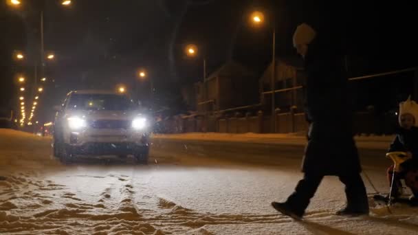 Mama zabiera dzieci na sanki, snowcat po drugiej stronie ulicy w zimie w nocy podczas opadów śniegu. Reflektory samochodu oświetlają drogę — Wideo stockowe