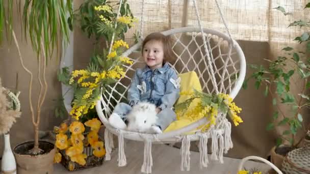 Küçük kız hasır bir salıncakta sallanıyor ve neşeli bir Paskalya gününde beyaz bir tavşanı okşuyor. — Stok video