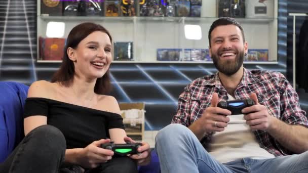Jovem casal jogando videogames, sentado em cadeiras em um clube de jogos com controladores em suas mãos — Vídeo de Stock