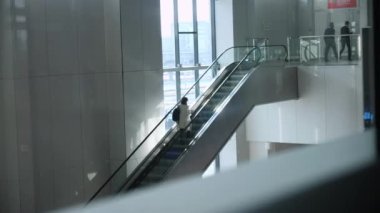 Sırt çantalı bir kadın binada yürüyen merdivene çıkıyor.