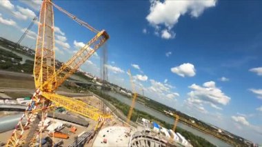 Sarı kule vinç inşaat alanındaki beton stadyum binasının yakınındaki yük asansörlerini kaldırıyor. Hava kamerası dönüyor ve iniyor.