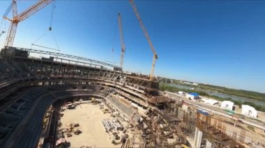 Metal çatı iskeleti, güneşli bir günde şehrin semtindeki spor stadyumunun inşaat sahasında vinçlere asılıyor.