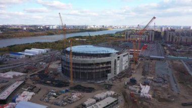 Büyük spor stadyum binası ve durgun nehir kıyısındaki vinçler yaz günü hava manzaralı bulutlar altında.