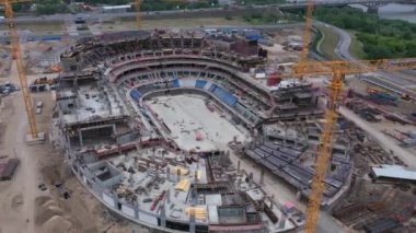 Oval spor arena binası ve kule vinçleri şehir nehir kenarı hava manzaralı inşaat sahasında. Sanayi iş akışı